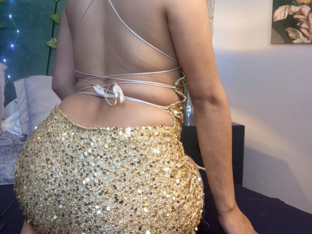 EmmaaRodriguez's ass in a gold, sequined skirt with white straps on her back, sitting on a bed, showing her tan skin and toned arms.