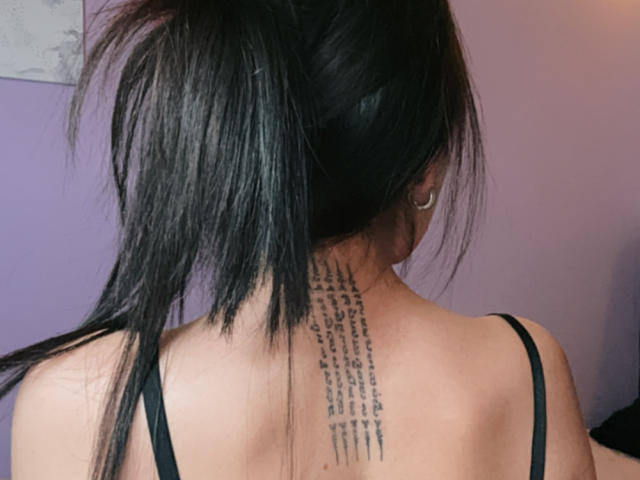 IzzysMisss has black tattooed text running vertically down her back, above her black strap top, with long black hair in a ponytail, and a small hoop earring.