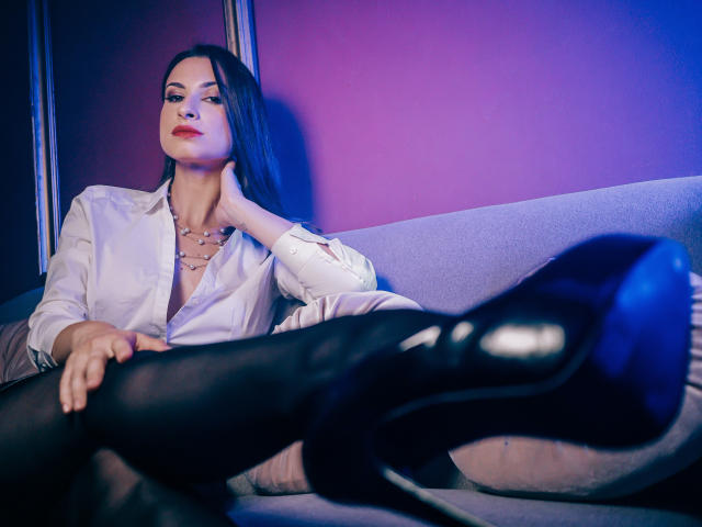 QueenSerenne, met lang zwart haar, een wit shirt, zwarte panty's en zwarte hoge hakken, ontspannen met een hand op haar nek, benen gekruist, in een kamer met blauw en paars licht..