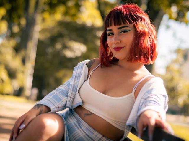 KarinaBlake met een rode bob, een witte top, een blauw geruit shirt en hoge shorts, zittend buiten, lachend, met tatoeages zichtbaar en rode lippenstift, met een ontspannen en zelfverzekerde uitstraling..