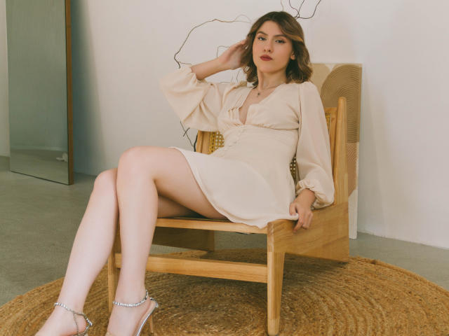 MaryBess with wavy brown hair, pale skin, and a cute face, wearing a beige dress and silver heels, sitting with legs crossed, hand on head, on a wooden chair.
