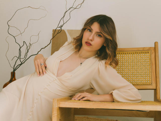 LizzieRossa with wavy brown hair, black bra, and tattoo on shoulder, looking over shoulder, hand in hair, showing her tits and cute face.