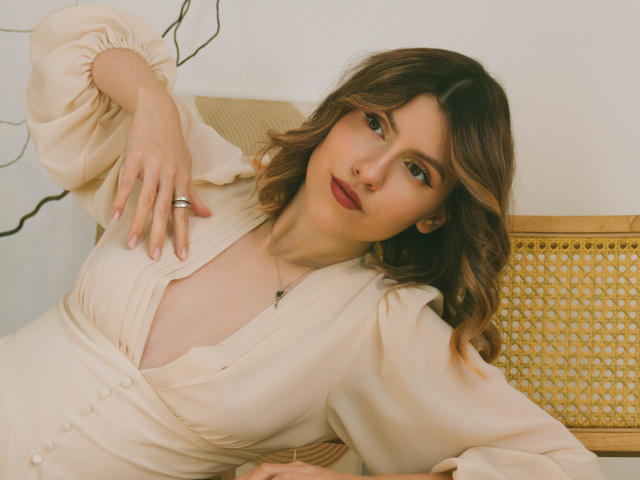 MaryBess with wavy brown hair, red lipstick, and a beige dress, lying on a yellow wicker chair, hand on chest, showing her tits, ring on finger, soft expression.