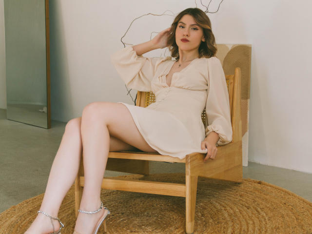 MoollyEvanns, topless, wearing black lace garter, white thong, black polka-dot stockings, black high heels, crouched, showing her ass, red hair, white chair, marble-patterned wall.