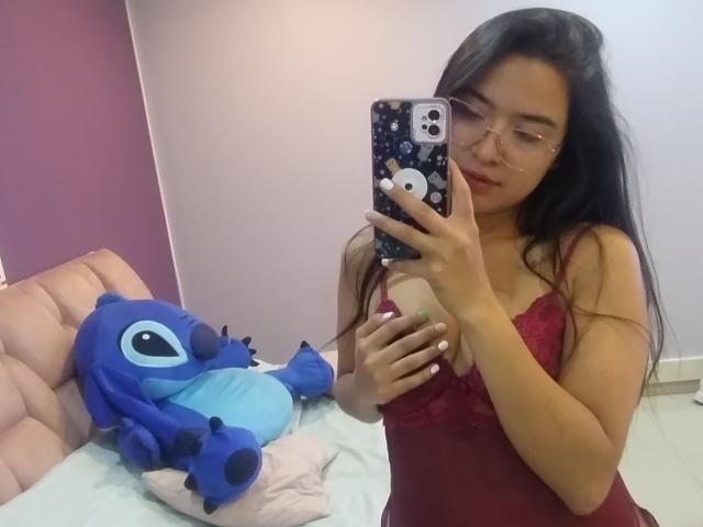 KeylaMeins, con i suoi lunghi capelli neri, indossa un abito intimo rosso in pizzo e si scatta un selfie allo specchio, tenendo un peluche Stitch blu su un letto rosa. Le tette di KeylaMeins sono visibili..