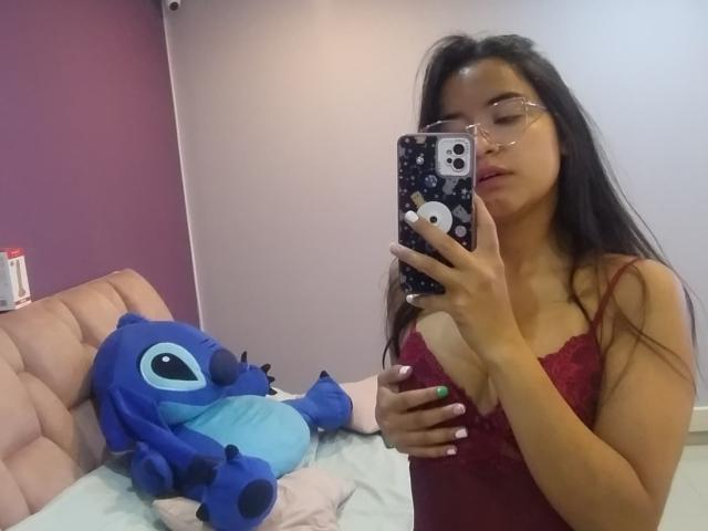 KeylaMeins with long black hair and glasses, wearing a red lace top, holds a phone with a blue patterned case, standing next to a blue Stitch plushie on a bed.