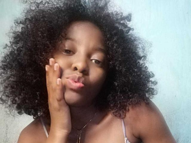 MarfSexy with dark curly hair, puffy lips making a kissy face, hand on cheek, wearing a strap bra, brown skin, looking slightly to the side, against a light background.