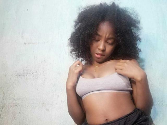 MarfSexy with curly black hair, wearing a grey sports bra, pulling down black pants, showing her small tits and slightly visible pussy, looking down with a serious expression.
