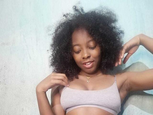 MarfSexy has dark curly hair, brown skin, small tits in a light gray bra, and a gold necklace, looking down with a slight smile. She's raising one arm.