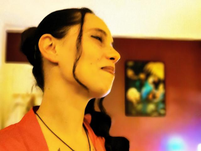 Nanastazia with dark hair in a braid, yellow light on her face, wearing a red shirt, necklace, and a small mole under her right eye, looking to the side.