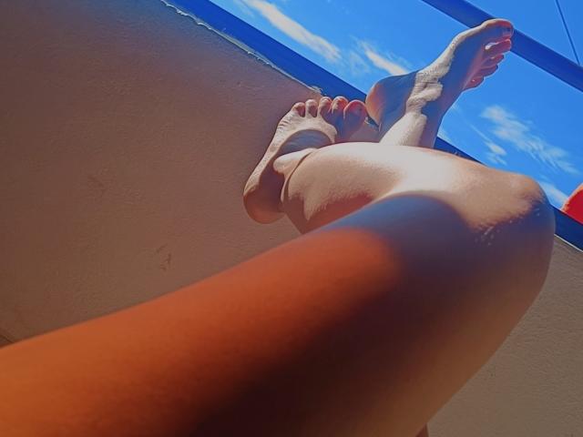 JulianaTasty's legs and feet are raised, showing her bare soles and red-painted toes against a blue sky and white wall. Her thighs are prominent in the foreground.