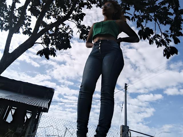 JulianaTasty with tall, slim, blue-jean-clad legs, green crop top, and glasses, standing under a tree, looking up, with a bright, cloudy sky and a small building in background.