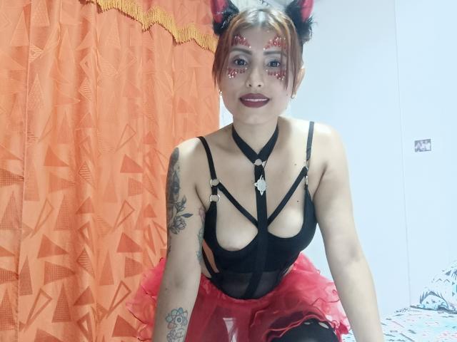 JulianaTasty with cat ears, red face paint, black strappy top, red tutu, tattoos, and red lipstick, kneeling on a bed, showing her tits and small ass.