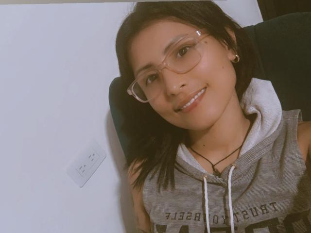 JulianaTasty with glasses, smiling, wearing a grey sleeveless hoodie, black hair, and small earrings, sitting in a dark chair. Her cute face and small nose are visible.