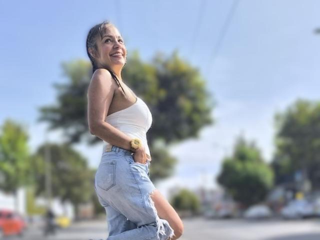 LailaCastillo, com seios pequenos, vestindo uma camiseta branca e jeans azul claro, sorrindo, perfil lateral, relógio no pulso, ao ar livre, ensolarado, árvores verdes ao fundo, olhando para cima..