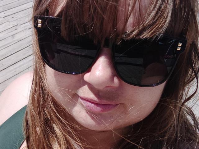 DorothySummer has light brown hair, wearing black sunglasses with gold accents, and a green top, showing a slight smile with pink lips. Her nose is visible, with light skin.