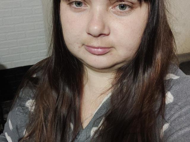 DorothySummer has pale skin, green eyes, and brown hair with bangs, wearing a gray, white-patterned top, with a slight, shiny pink lip. Her face is centered.