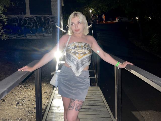 PamellaSweet with blonde hair, strapless top showing her tits, gray skirt, tattoos on her thigh, standing on a wooden bridge at night, arms on railings, glowing flash in front.