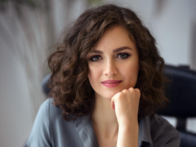 StaceyNetty with curly brown hair, blue eyes, and pink lipstick, resting her chin on her hand, wearing a gray shirt, looking directly at the camera with a slight smile.
