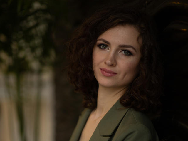 StaceyNetty has curly brown hair, green eyes, and a slight smile. She's wearing a green jacket, and her ass isn't visible. She looks cute and confident.