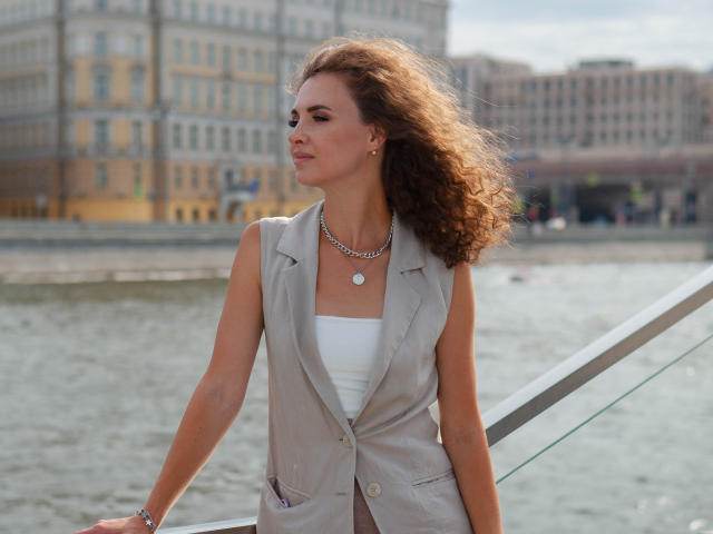 StaceyNetty with curly brown hair, wearing a gray sleeveless vest over a white tube top, silver necklace, and bracelet, leaning on a glass railing by a waterway.