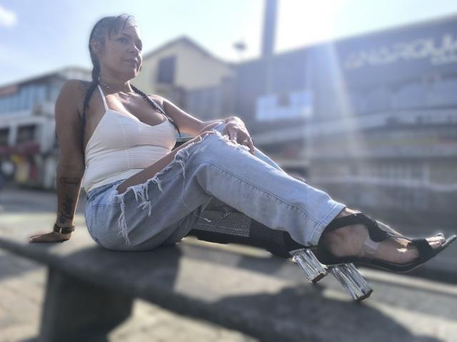 Laila Castillo, con i capelli intrecciati, una canotta bianca e jeans strappati, seduta su una panchina, indossa tacchi a piattaforma trasparenti, la luce del sole crea riflessi, ha un tatuaggio sull'avambraccio destro, e appare sicura di sé..