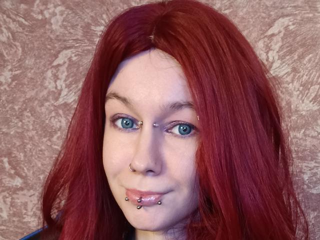 Vermilia with red hair, blue eyes, nose ring, and lip piercings, smiling slightly. She has a septum piercing and lower lip piercing. Her skin is fair.