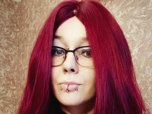 Vermilia with red hair, glasses, nose piercing, and lower lip piercing, looking at camera, slight smile, light skin, textured wall in background.