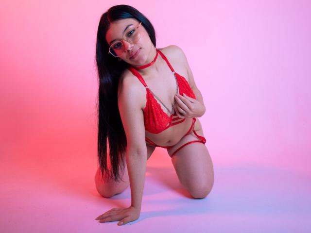 Ebba with long black hair, wearing red lace bra and thong, kneeling on white floor, hand on chest, red choker, glasses, pink and blue lighting, looking at camera.