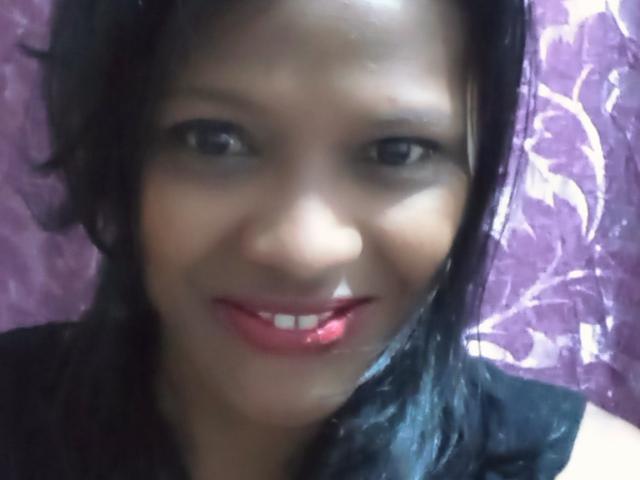 Irdah has dark brown skin, black hair, and is smiling with red lipstick. Her small tits are slightly visible under a black top. The photo is blurry.