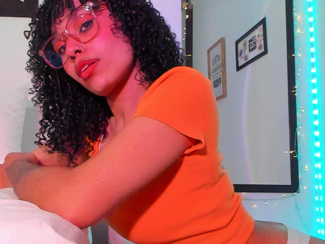 NickiBloom with curly black hair, red lipstick, and glasses, wearing an orange top, leaning on a bed, looking at the camera with a slight smile. Blue LED lights.