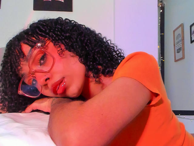 NickiBloom with curly black hair, red lipstick, and orange glasses, wearing an orange shirt, lying on a bed, resting her head on her hand, looking at the camera.