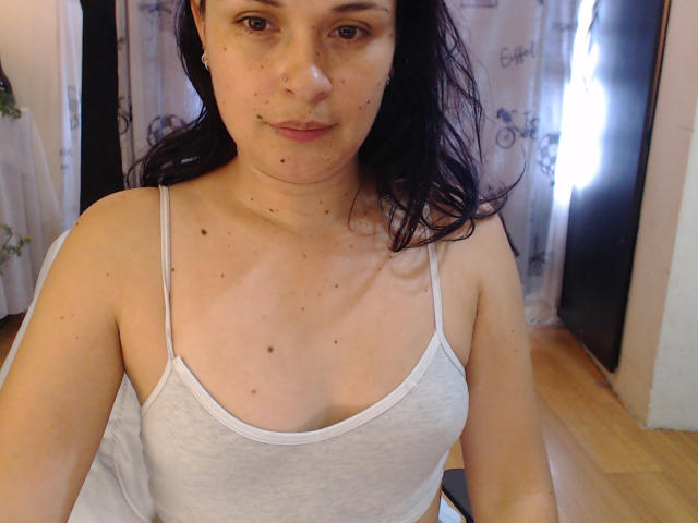 WendySchulz has light skin, small brown eyes, and dark hair. She's wearing a grey tank top, showing her small tits and some freckles and moles on her chest.