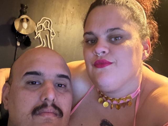 YvannaHetDreammix with pink lipstick, nose ring, and gold necklace, her large tits visible, stands next to bald, mustached dude, both smiling. Neon wall art in background.