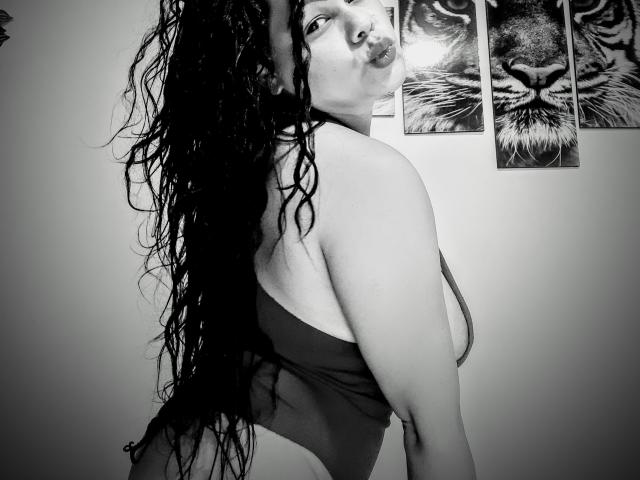 Black-and-white photo of AlhelQueen with wet, curly hair, pouty lips, and a revealing one-piece that shows her big ass and sideboob. Tiger paintings in the background.