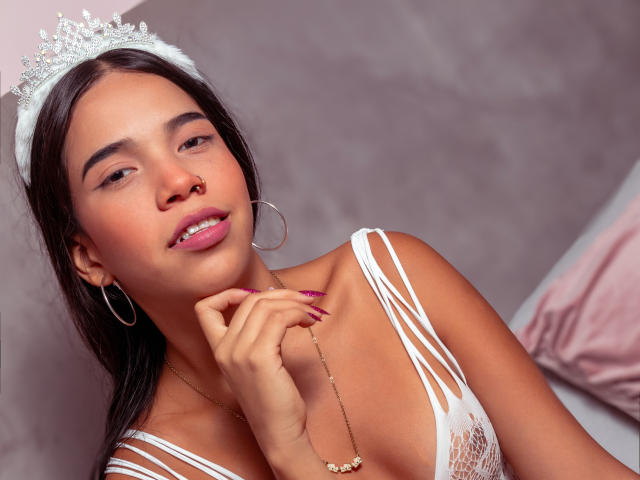 Mary Smith, com cabelo castanho, usando uma tiara prateada, brincos de argola e uma blusa branca com alças finas, mostrando seu batom rosa e segurando o queixo, com um colar..
