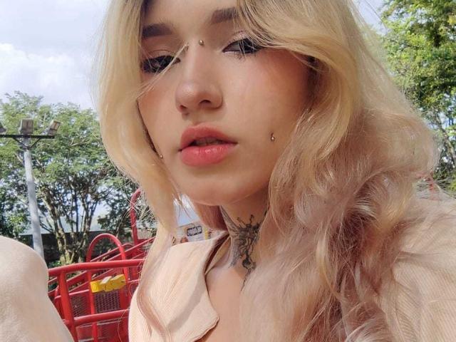 CharlotteAndLiam with blonde wavy hair, nose piercings, lip piercings, tattoos, pink lipstick, and a soft pink top, outdoors with red metal chairs in the background.