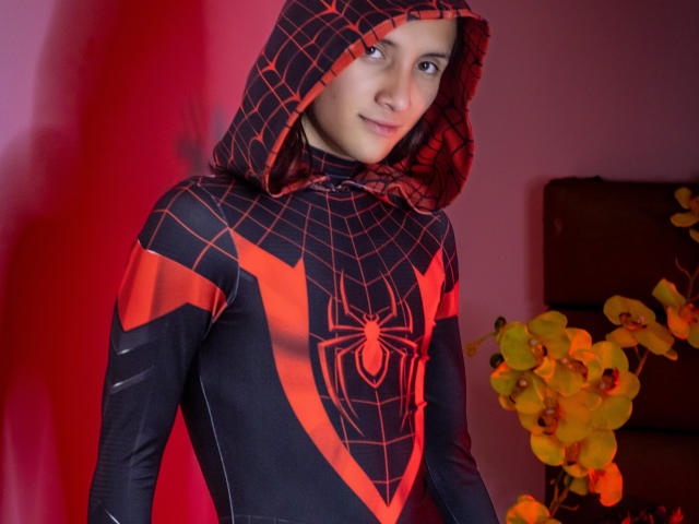 FelixNelson indossa una felpa di Spider-Man, rossa e nera, con un simbolo di ragno. FelixNelson ha un sorriso carino, occhi marroni e capelli castani corti. Sullo sfondo, ci sono orchidee arancioni..