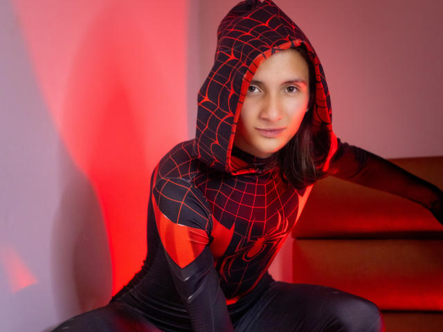 FelixNelson, gekleed in een Spider-Man hoodie met rode webpatronen, hurkt neer en toont een schattende glimlach, terwijl rood licht een aantrekkelijke gloed op hun gezicht werpt..