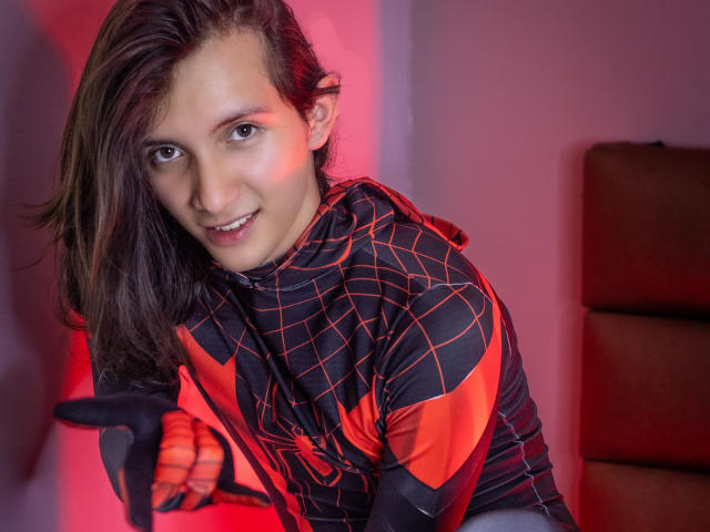 FelixNelson, gekleed in een zwart en rood Spider-Man kostuum, heeft bruin haar dat tot op zijn schouders reikt, en glimlacht met een speelse uitdrukking, terwijl hij een zwart rekwisit vasthoudt in een kamer met rood licht..