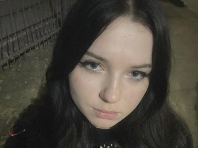 ToriFran, con piel pálida, cabello oscuro y ondulado, y ojos azules, tiene una expresión de labios fruncidos y viste ropa negra, sin que se vean sus senos. Está mirando directamente a la cámara..