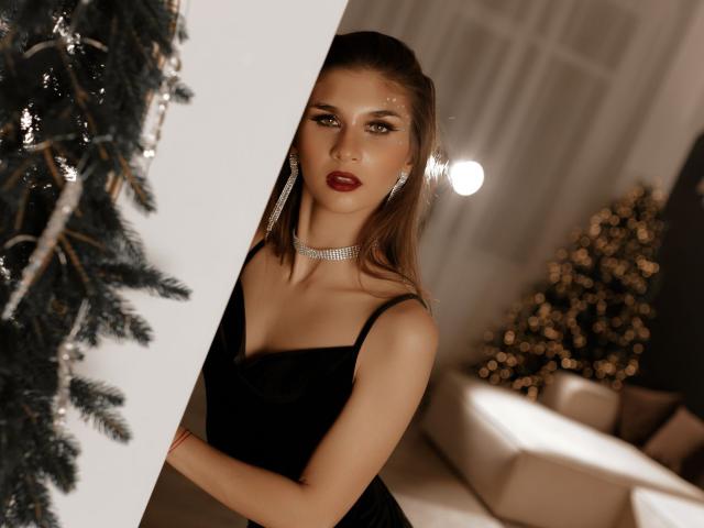MiaAxel with brown hair, red lipstick, and black dress, wearing a sparkly choker, peeks from behind a Christmas tree, looking sexy with tits visible in the foreground.