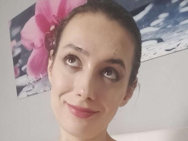 JuliaHuntis with shiny skin, brown eyes, and pink lipstick, smiling slightly, has a pink hibiscus flower in her dark hair, and a blue and white background.
