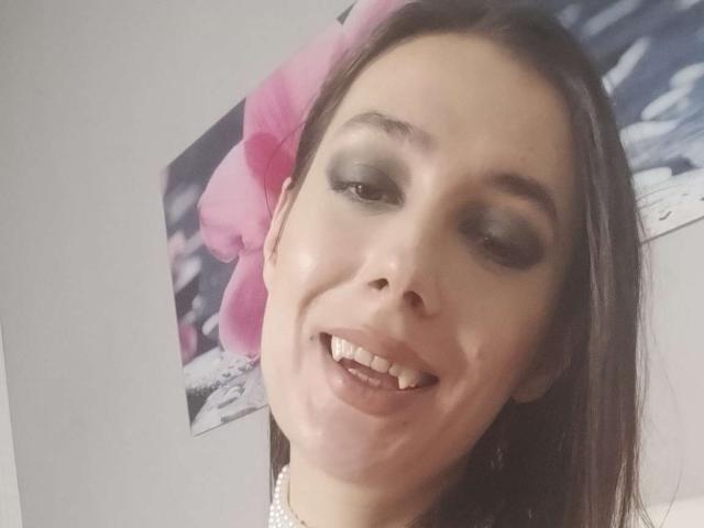 JuliaHuntis with dark brown hair, pink flower, and makeup, smiling with visible teeth, gray eyes, and white necklace, in a selfie. Her face is slightly tilted to the right.
