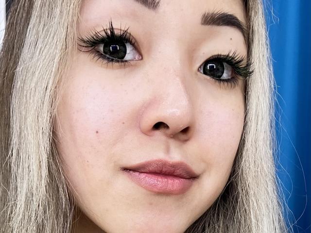 RenyLime's face, showing light brown skin, blonde hair, thick black eyelashes, small nose, pink lips, and slightly parted mouth, with a blue background blur.