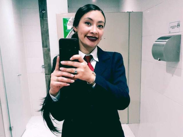 TatianaRiosLove with long black hair, dark lipstick, in a blue suit, white shirt, red tie, taking a selfie in a bathroom, holding a black phone.