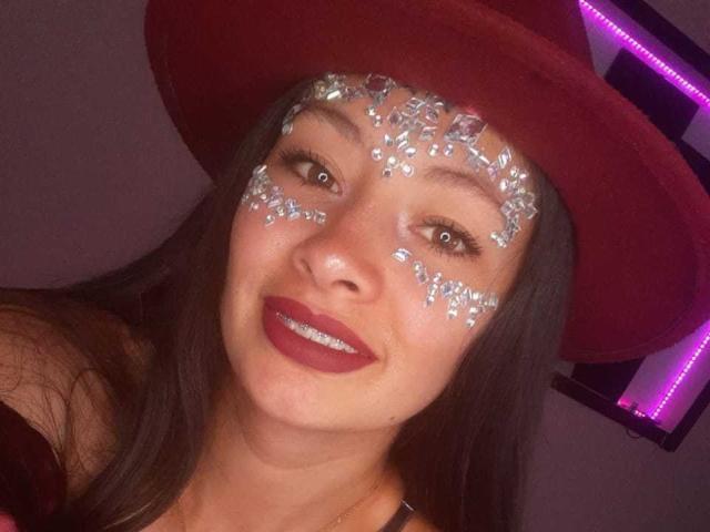 TatianaRiosLove with dark brown hair, wearing a red hat, has glitter on her forehead and cheeks, red lipstick, and a smile, with visible eyelashes and a gold necklace.