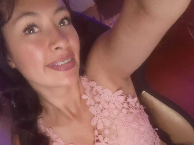 TatianaRiosLove with brown eyes, smiling, wearing pink lace top, arm raised, showing her tits, brunette hair, purple lipstick, and slight blush, in a selfie.