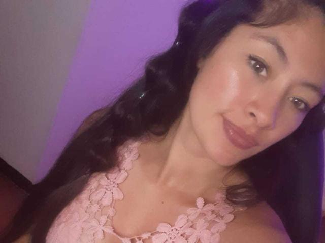 TatianaRiosLove has light brown skin, wavy black hair, and is wearing a pink lace top, showing her small boobs and subtle smile in a softly lit photo.