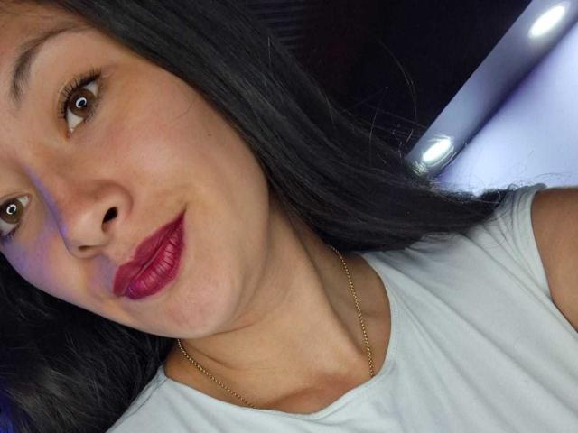 TatianaRios: Uma mulher de cabelos escuros, olhos castanhos e batom vermelho, vestindo uma camisa branca e um colar de ouro, sorrindo levemente. Seu rosto está centralizado na foto..
